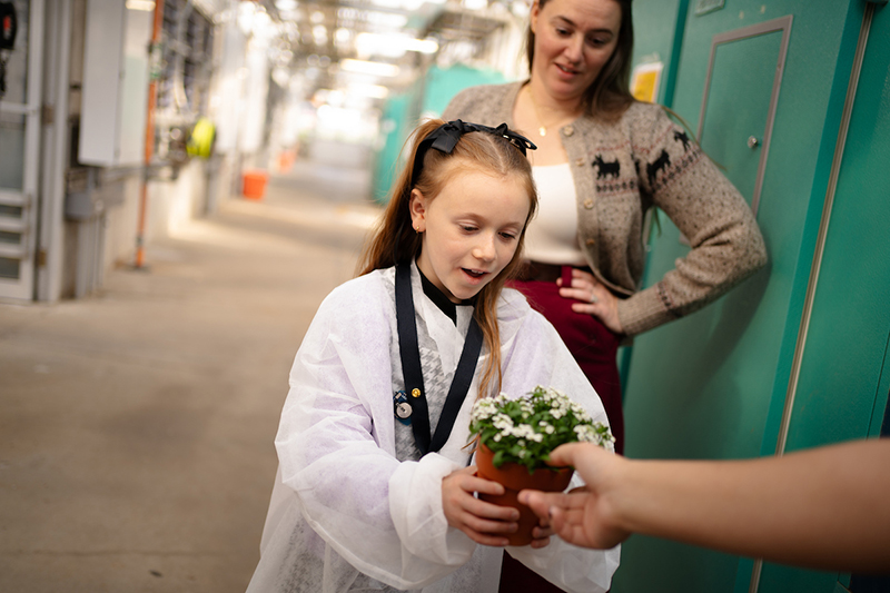 Girl in a lab coat receiving a small potted plant from someone while an adult stands behind her